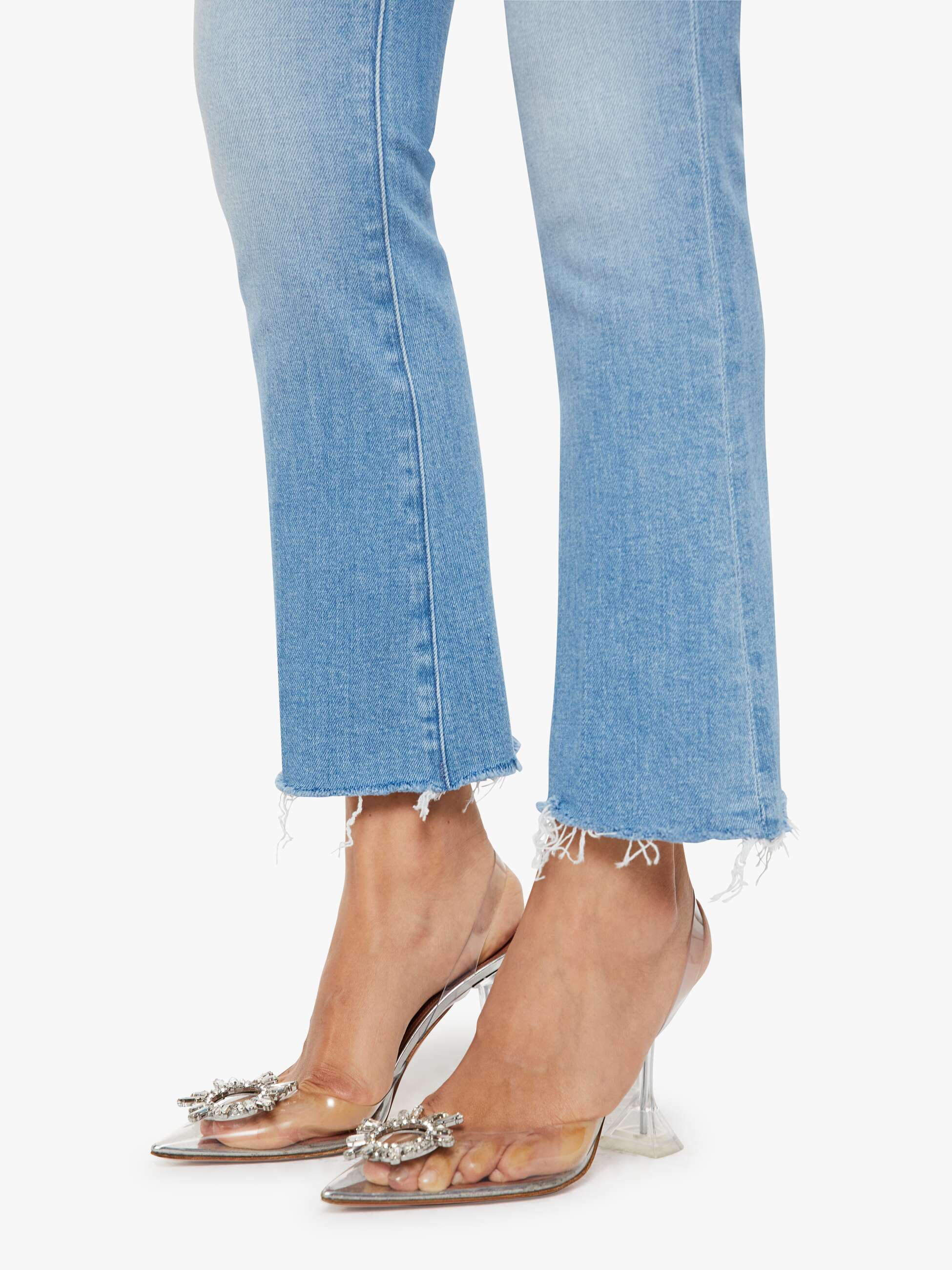 Close-up of light blue jeans with a frayed hem and clear high-heeled shoes on a white background