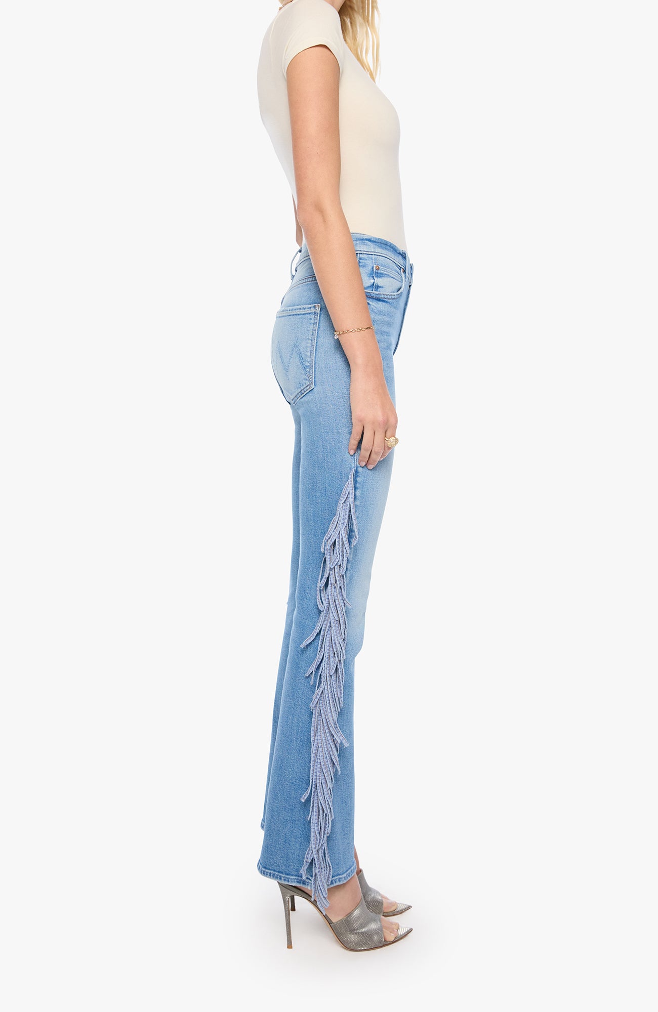 A side-profile view of a model wearing light-wash blue denim jeans with long fringe detailing running down the outer side of the legs, paired with a cream short-sleeve top and metallic gray high-heeled sandals against a plain white background.