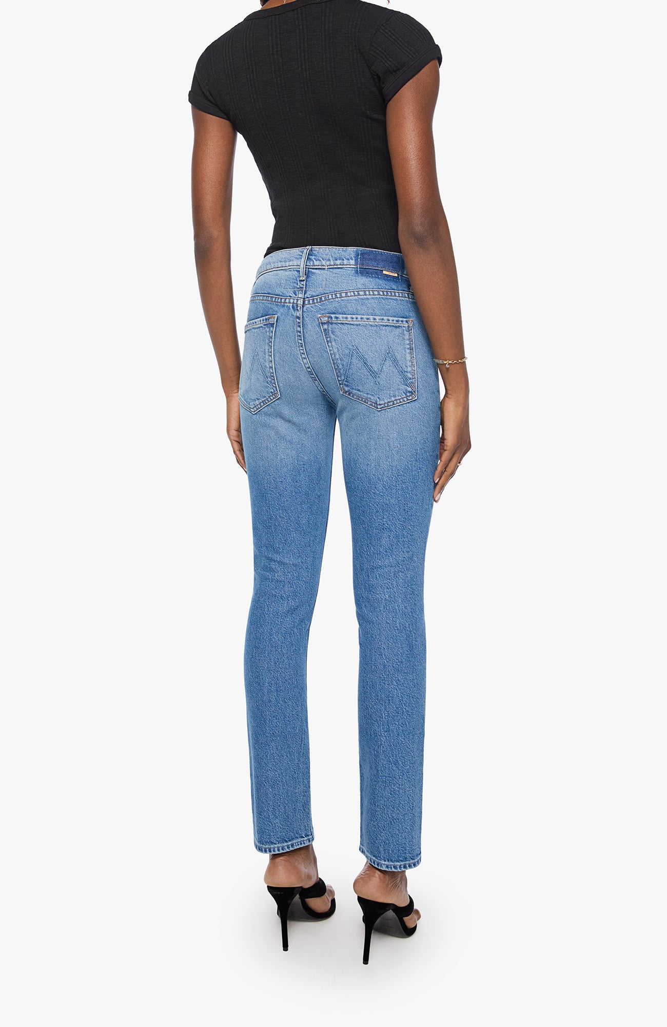A model shown from the back wearing light-wash blue Mary Jane jeans, a black short-sleeve top, and black high-heeled sandals against a light gray background.