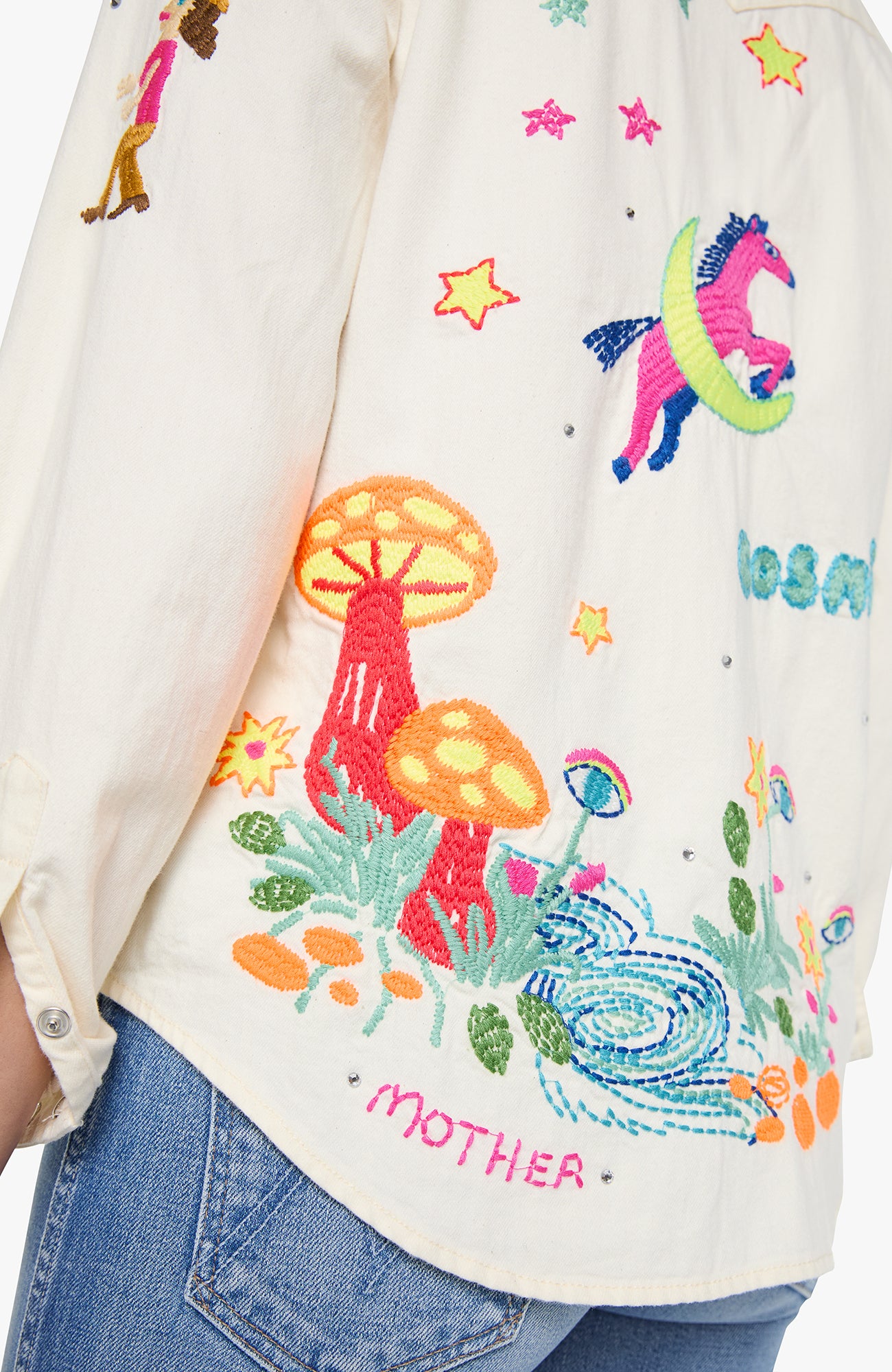 Close-up of a cream-colored embroidered shirt worn with blue jeans. The shirt features colorful embroidery of a pink unicorn on a crescent moon, orange and red mushrooms, stars, and the word 'MOTHER' in pink.