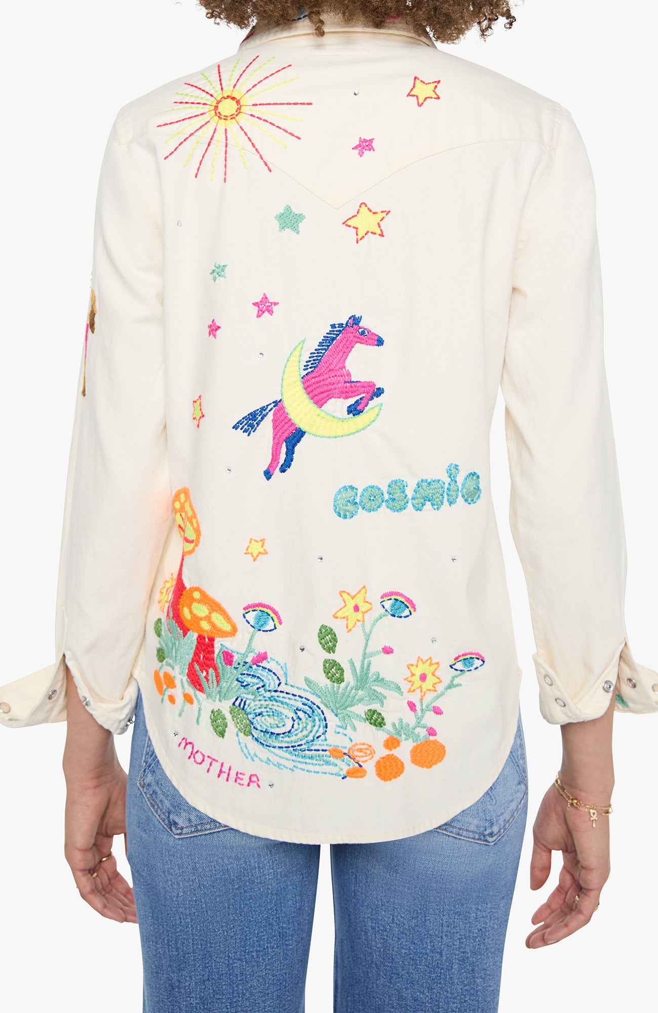 A woman shown from the back wearing a cream-colored long-sleeve button-up shirt with colorful embroidered designs, including a unicorn, stars, flowers, and the words 'cosmic' and 'MOTHER'. She is wearing blue jeans against a plain white background.