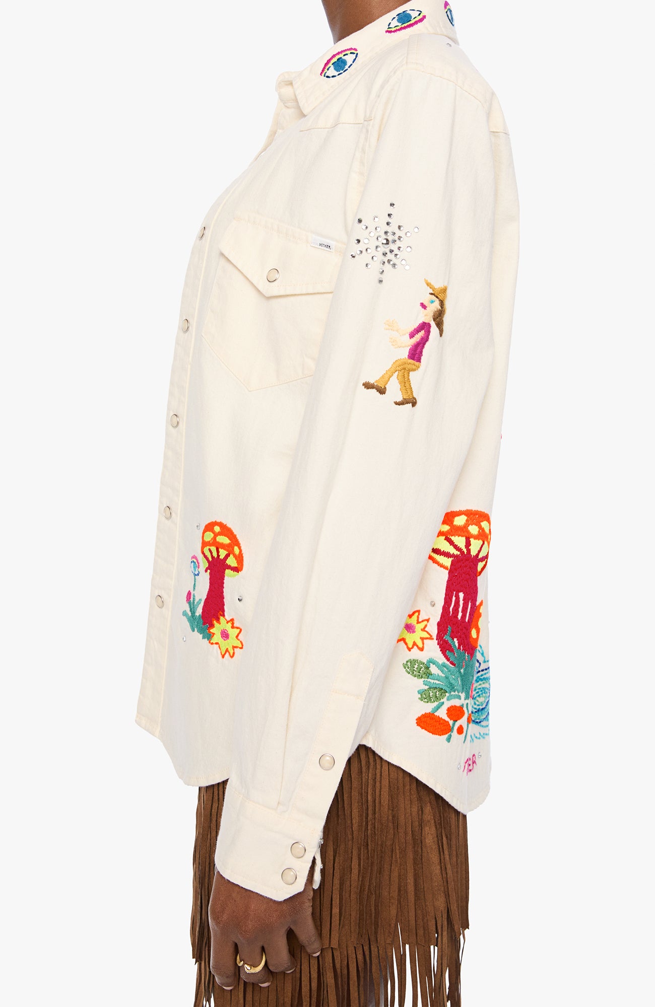 An off-white button-up shirt with colorful embroidered details, including mushrooms, flowers, and a small dancing figure, shown on a model wearing a brown fringe skirt against a light gray background.
