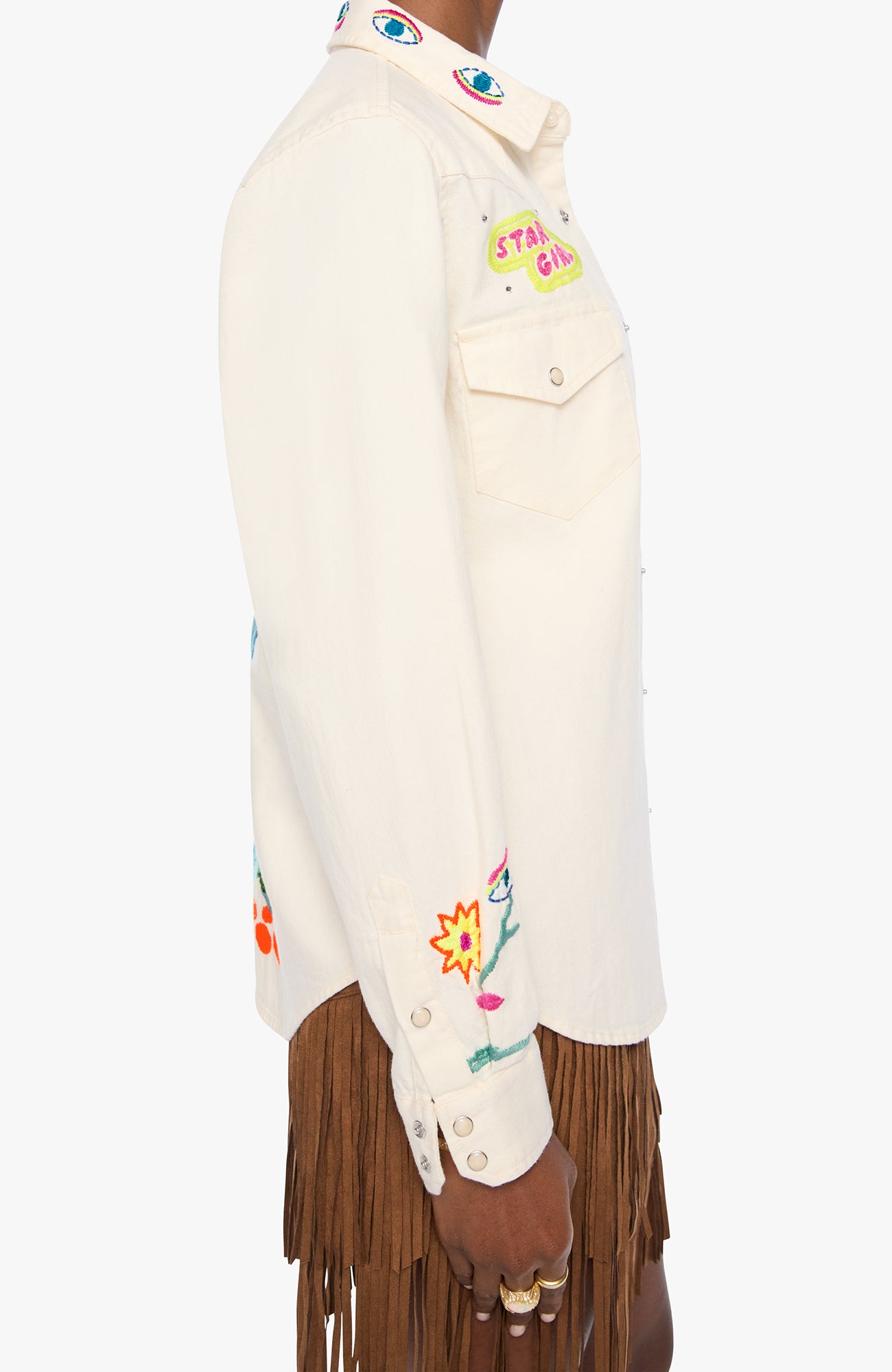 Side view of a model wearing a cream-colored denim jacket with colorful embroidered details, including a 'STAY GIRL' patch on the chest and floral embroidery on the sleeve, paired with brown fringe pants.