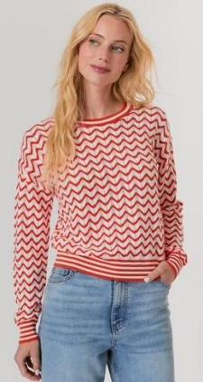 Woman wearing a red and white zigzag patterned sweater and blue jeans on a plain background