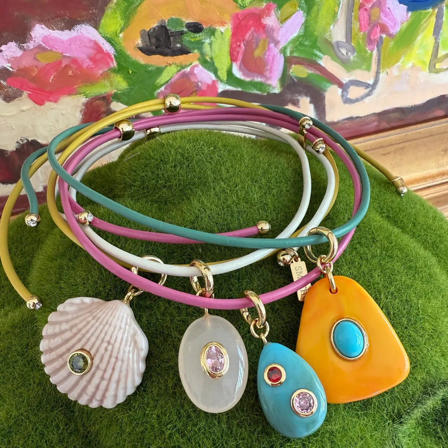 A stack of five multi-strand leather wrap bracelets with gold-tone hardware and colorful pendants, displayed on a green textured surface with a painted floral background. The pendants include a white seashell, a milky white oval with a pink stone, a turquoise teardrop with a pink stone, a bright turquoise oval, and a large orange teardrop with a turquoise center.