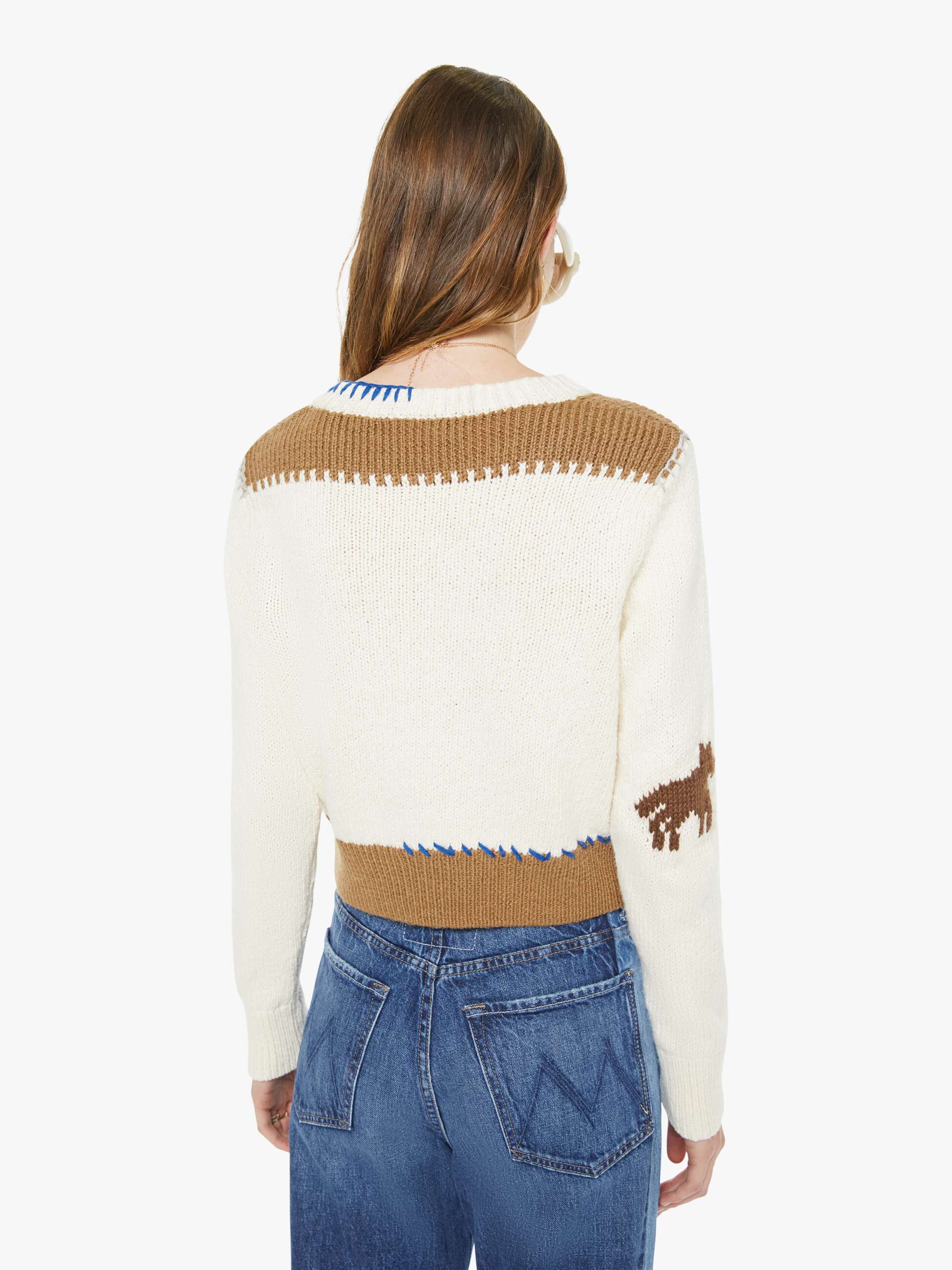 Person wearing a cream sweater with brown accents and blue jeans on a white background