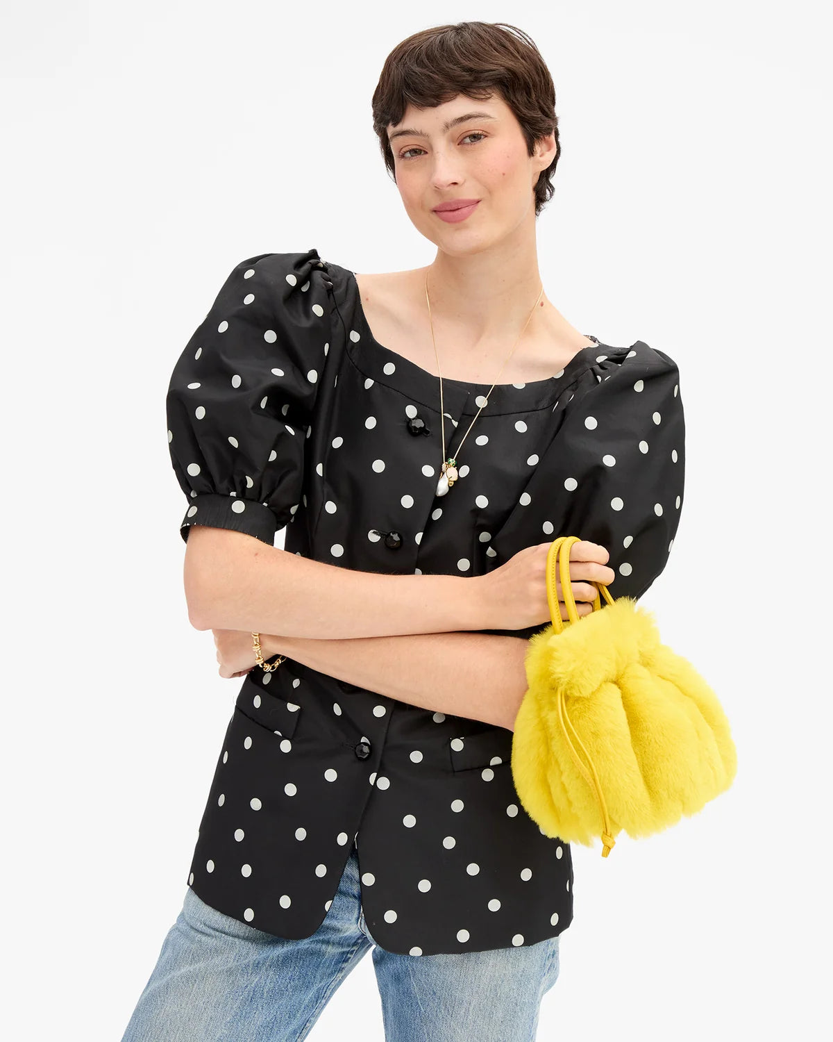 A model wearing a black puff-sleeve blouse with white polka dots, paired with light blue jeans and holding a bright yellow fuzzy handbag, set against a plain white studio background.