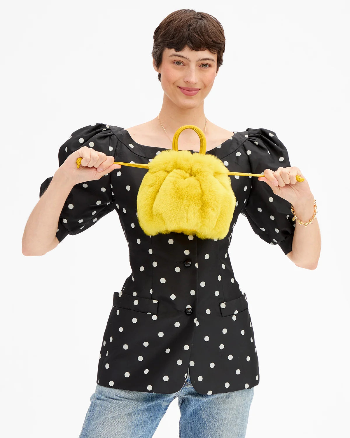 A model wearing a black puff-sleeve top with white polka dots holds up a bright yellow faux-fur handbag with a yellow top handle against a plain white background.