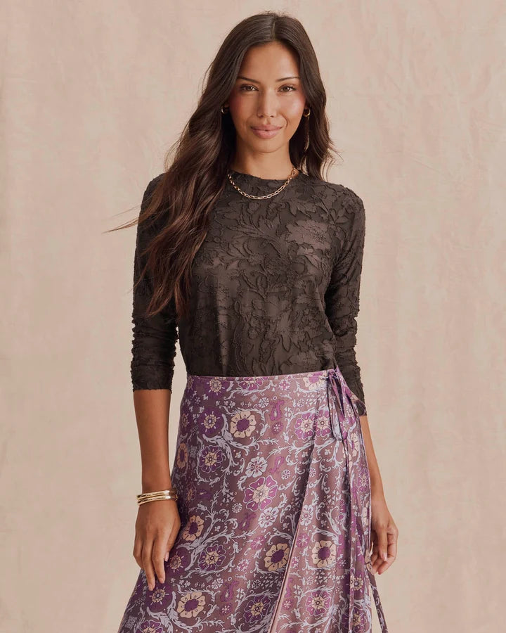 A woman modeling a dark brown lace long-sleeve top paired with a purple and pink paisley patterned wrap skirt, set against a warm beige textured background.