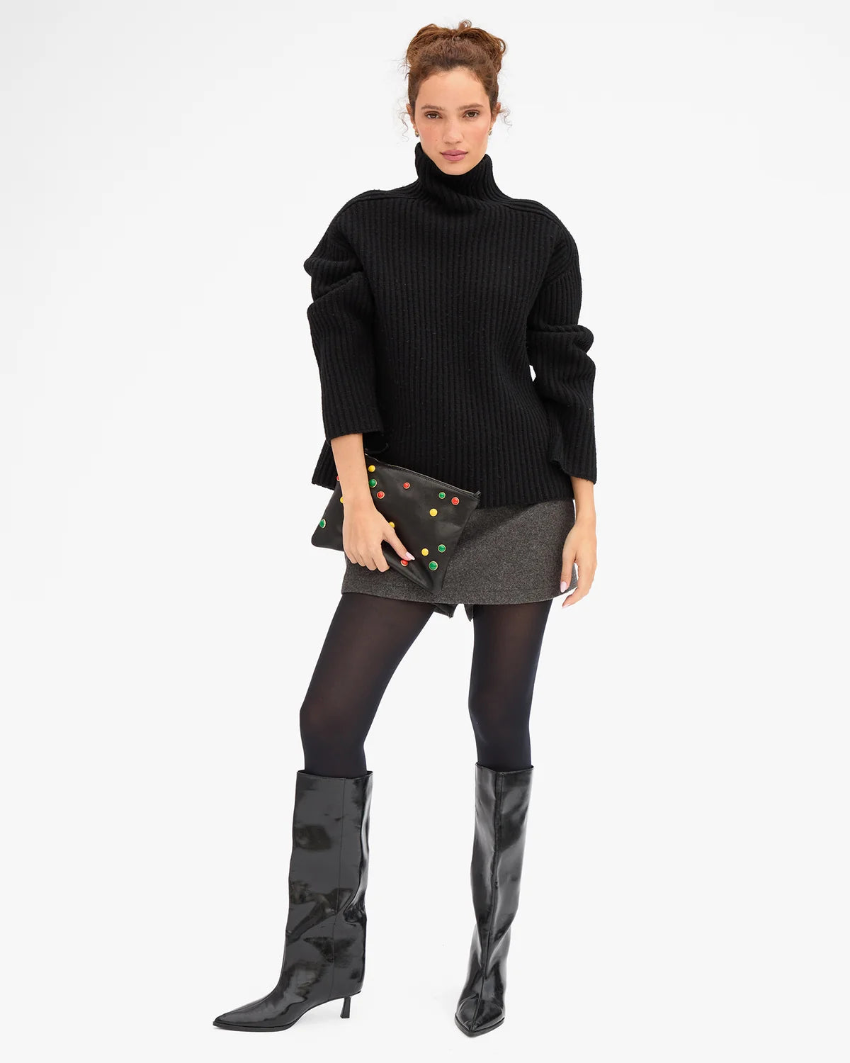 A model stands against a light gray studio background wearing a black ribbed turtleneck sweater, a gray mini skirt, sheer black tights, glossy black knee-high boots, and holding a black clutch decorated with colorful studs.