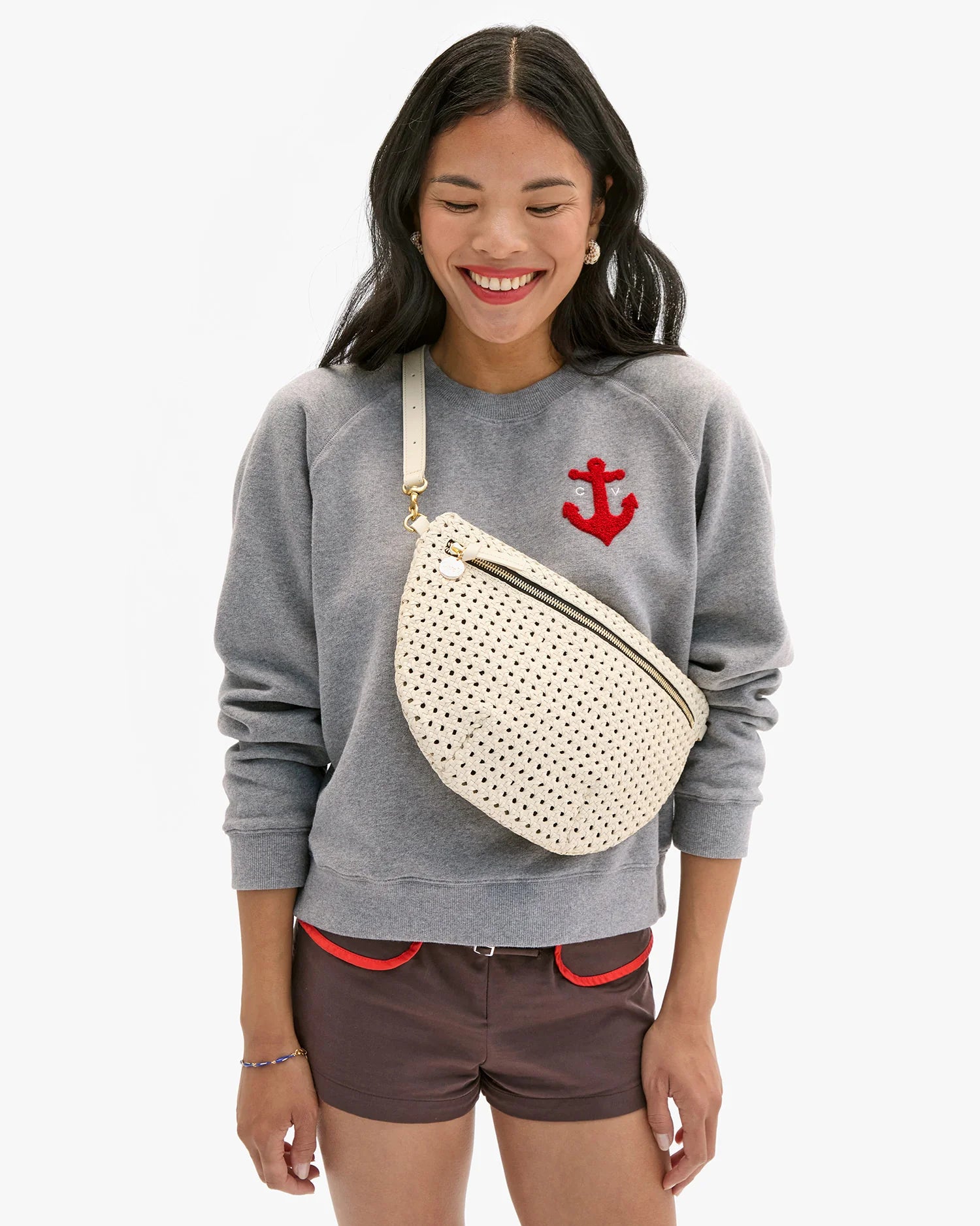 Woman wearing a gray sweatshirt with a red anchor design, brown shorts, and carrying a cream-colored perforated bag on a white background.