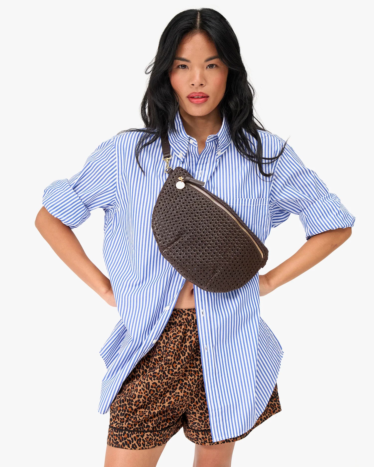 Woman wearing a blue striped shirt and leopard print shorts with a brown woven bag on a white background