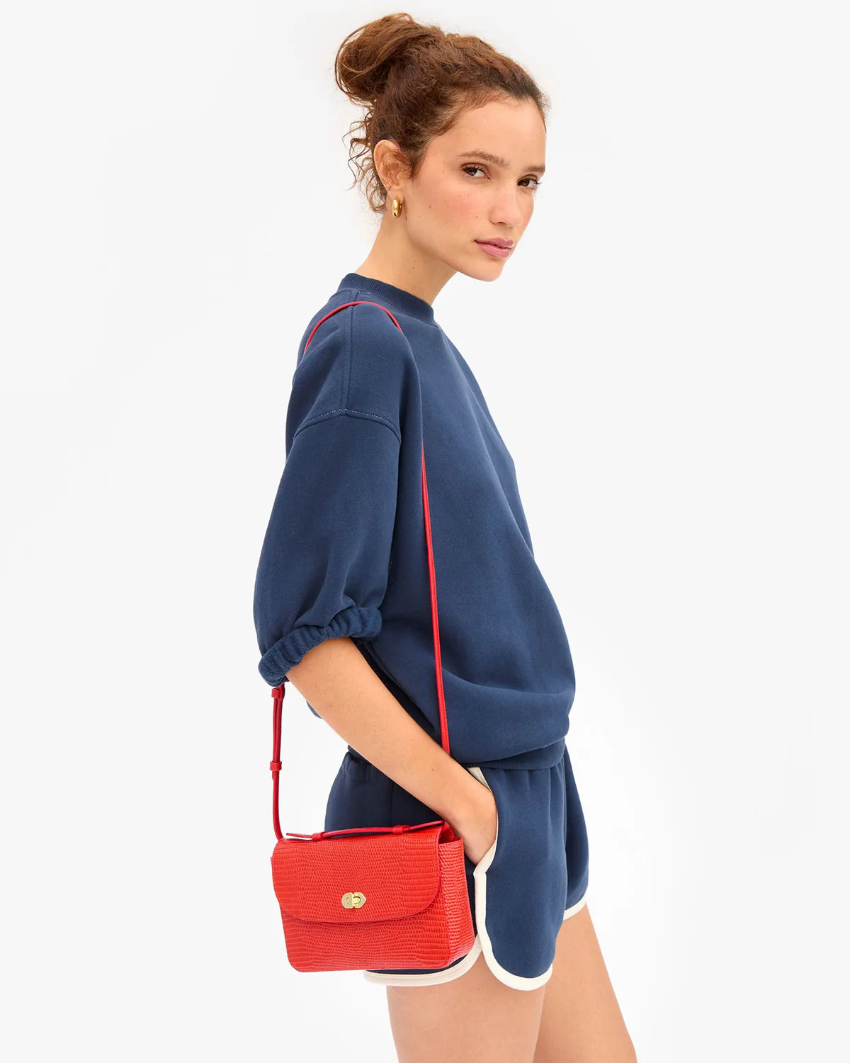 A woman modeling a navy blue sweatshirt and matching shorts, accessorized with a bright red textured crossbody bag with a gold clasp, against a plain white background.