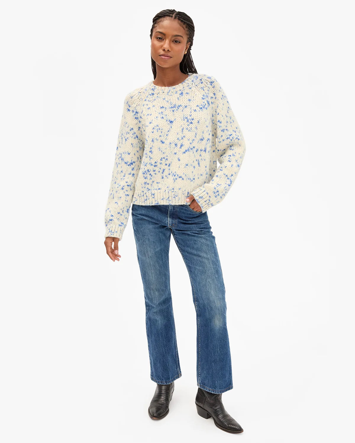 A model wearing a cream-colored knit sweater with a blue floral pattern, paired with blue jeans and black ankle boots, standing against a plain white studio background.