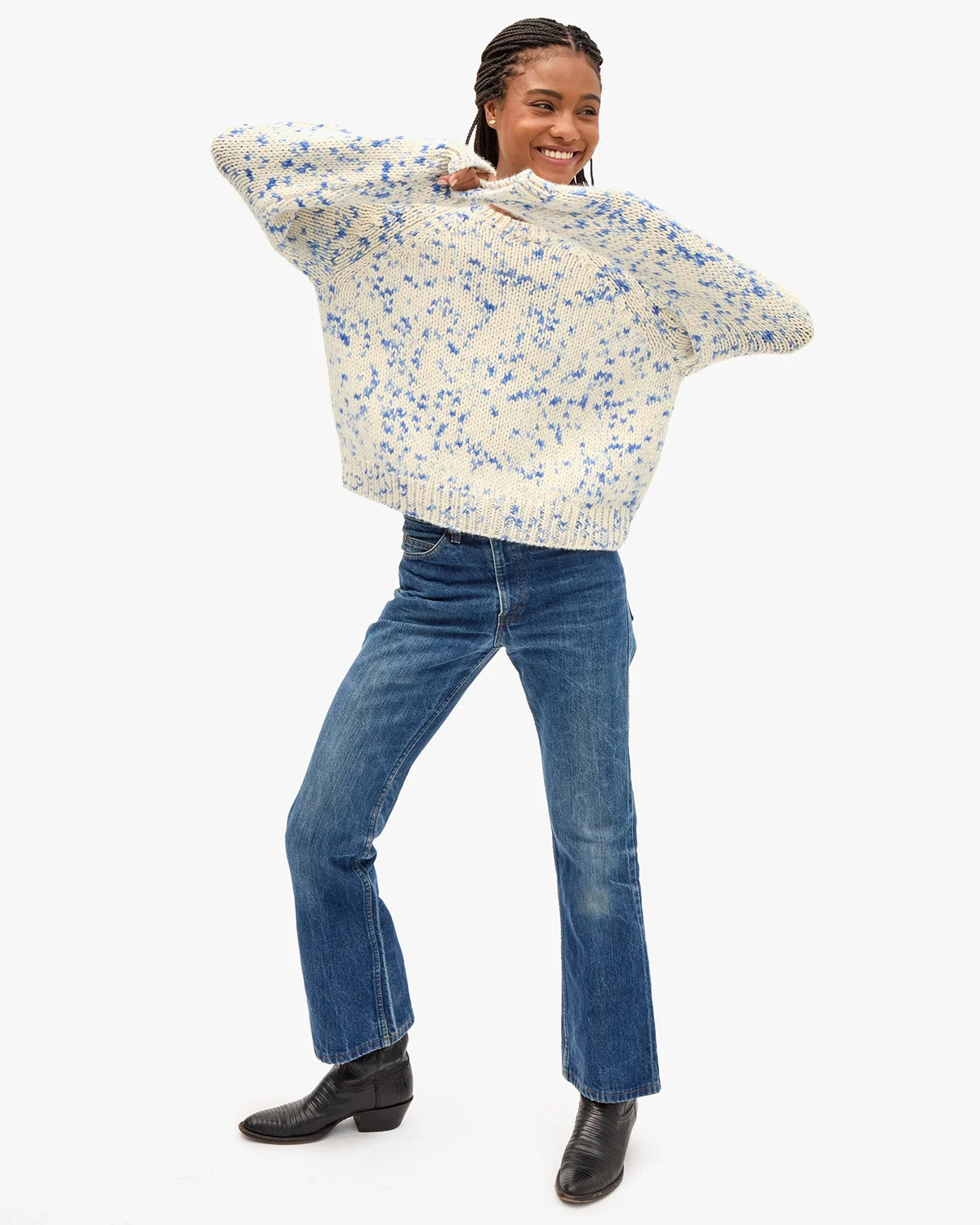 A woman modeling a cream knit sweater with a blue floral pattern, paired with blue jeans and black boots, against a plain white background.