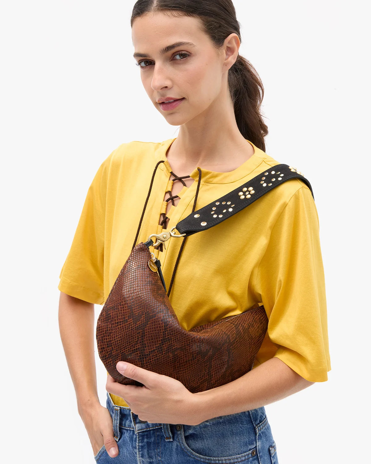 A person wearing a yellow top and blue jeans carries a brown snakeskin-patterned crescent shoulder bag with a black strap against a plain white background.