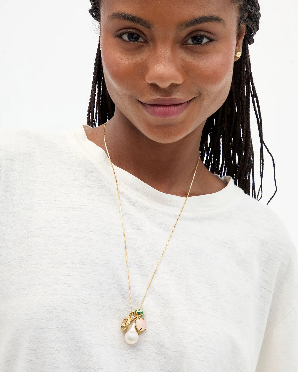A close-up of a model wearing a delicate gold chain necklace with a multi-piece pendant featuring a white pearl, a peach-colored bead, and small green and gold details, paired with a white crewneck top.