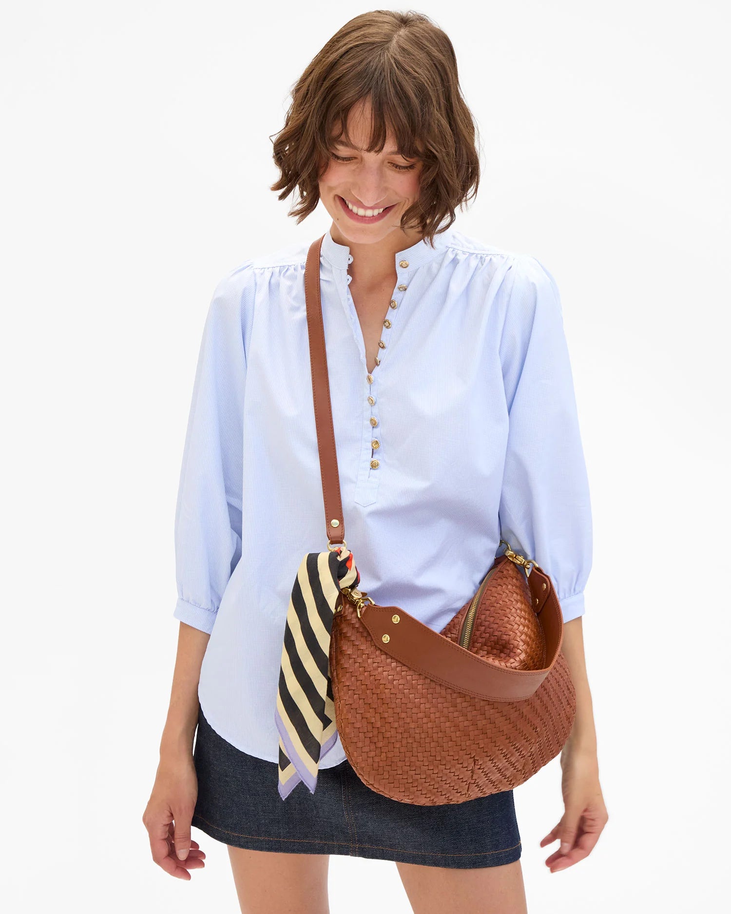 Woman holding a brown woven handbag with a striped scarf, wearing a light blue shirt and dark skirt on a white background