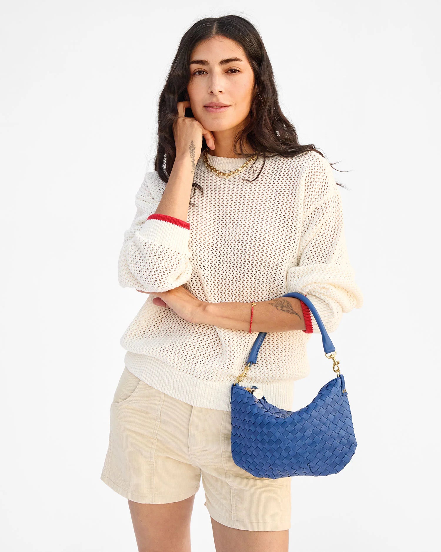 Woman wearing a white sweater and beige shorts, holding a blue handbag on a white background
