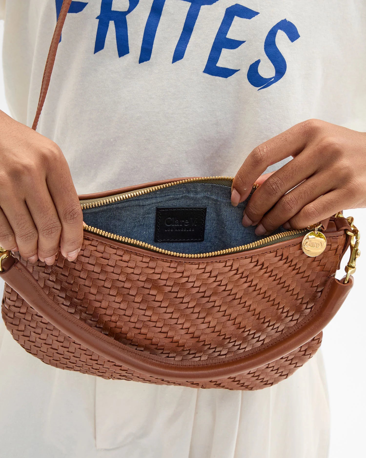 Brown woven handbag with gold zipper held by a person wearing a white shirt with blue text.