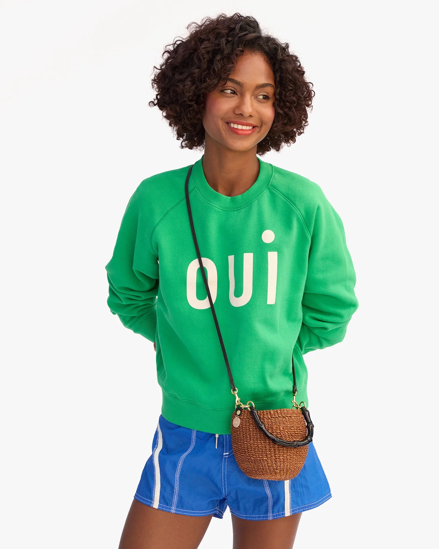 Woman wearing a green sweatshirt with 'QUI' text, blue shorts, and holding a brown bag on a white background