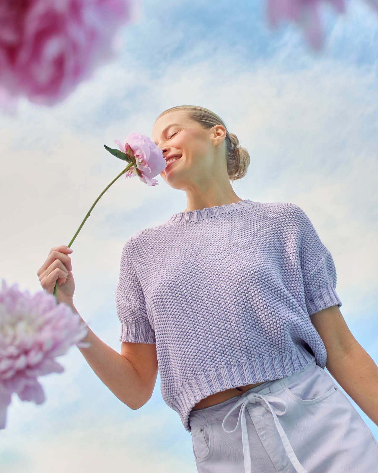 Woman in a light purple sweater holding a pink flower against a colorful sky background