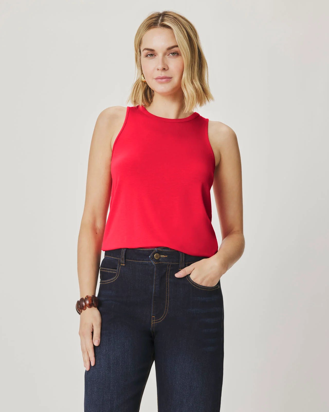 Woman wearing a red sleeveless top and dark jeans on a plain background