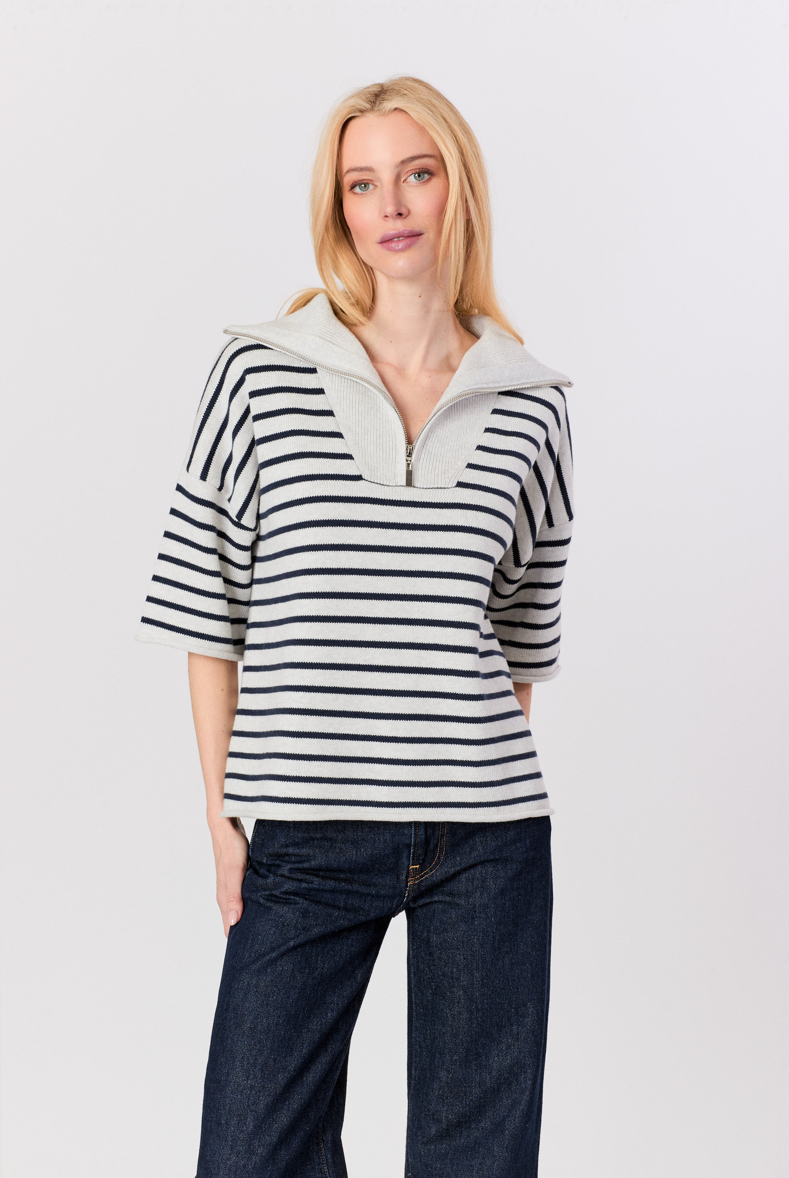A blonde woman wearing a navy and white horizontally striped knit top with a light gray mock neck collar and dark blue jeans, standing against a light gray studio background.