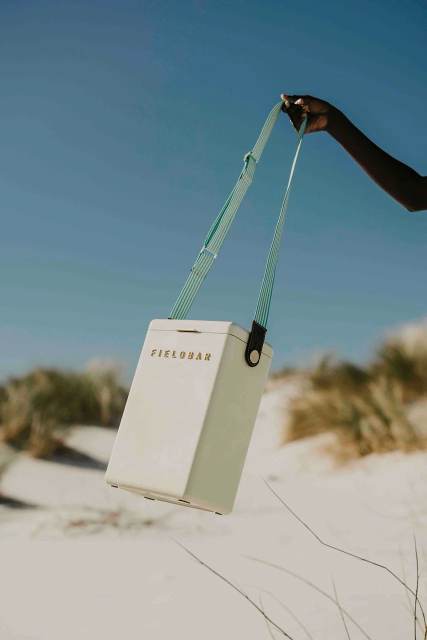 Person holding a beige cooler bag with green strap on a sandy beach.