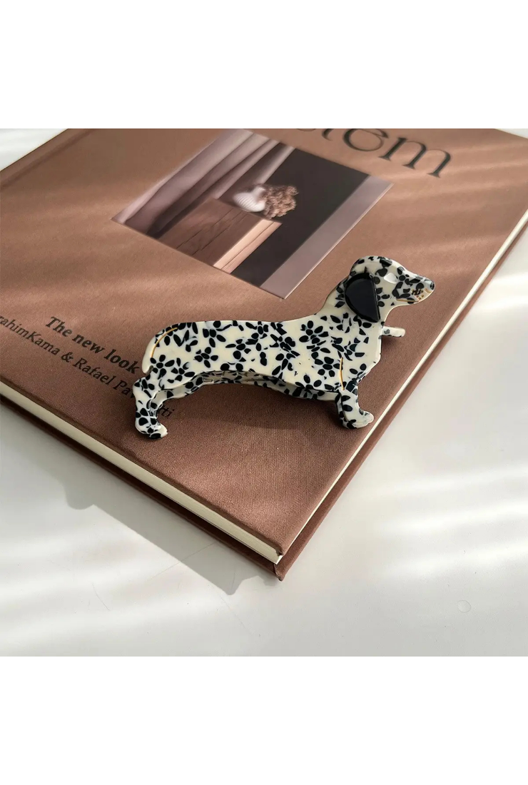 A small dachshund-shaped brooch with a black and white floral pattern, resting on the corner of a brown book.