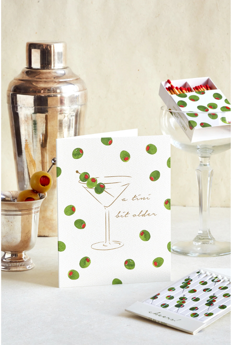 A white greeting card with a martini glass illustration and green-and-red olives, styled with a cocktail shaker, glass, matches, and a notebook on a light surface against a cream background.