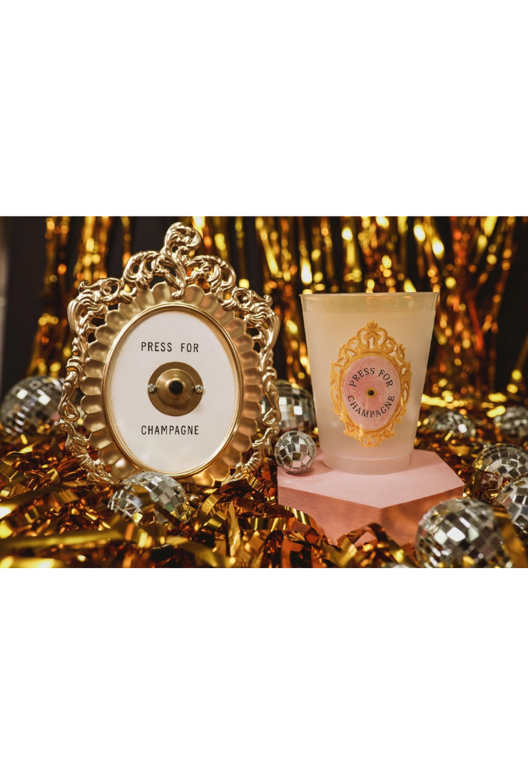 A champagne-themed decorative set featuring a gold ornate frame with a white dial labeled 'PRESS FOR CHAMPAIGE' and a frosted glass cup with a matching gold emblem on a pink pedestal, surrounded by gold tinsel and mirrored balls.