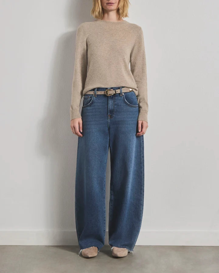 A model wearing a beige knit sweater, wide-leg blue jeans with a thin brown belt, and beige shoes, standing against a plain light gray wall.