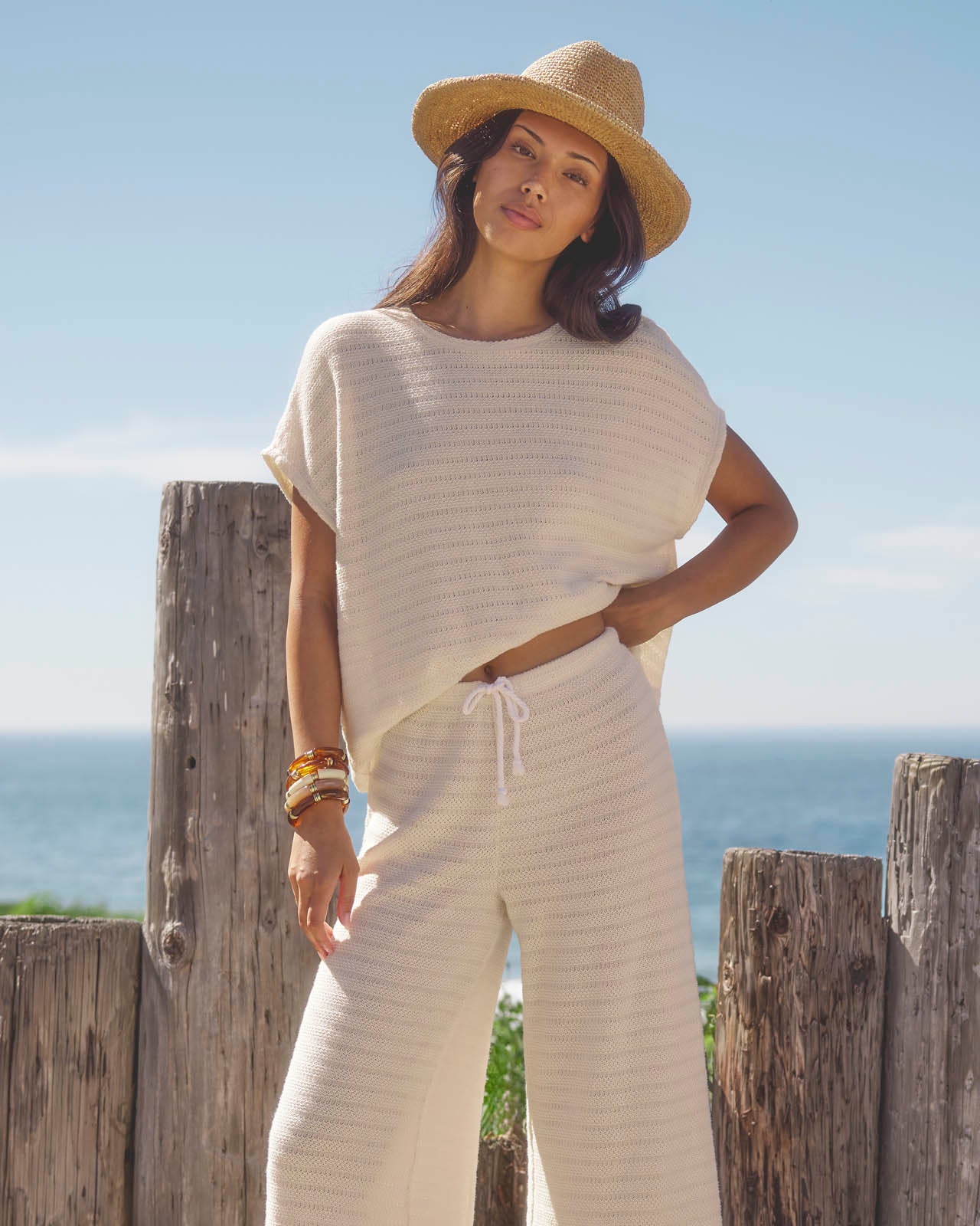 Woman in a white outfit and straw hat standing by a wooden post with ocean view
