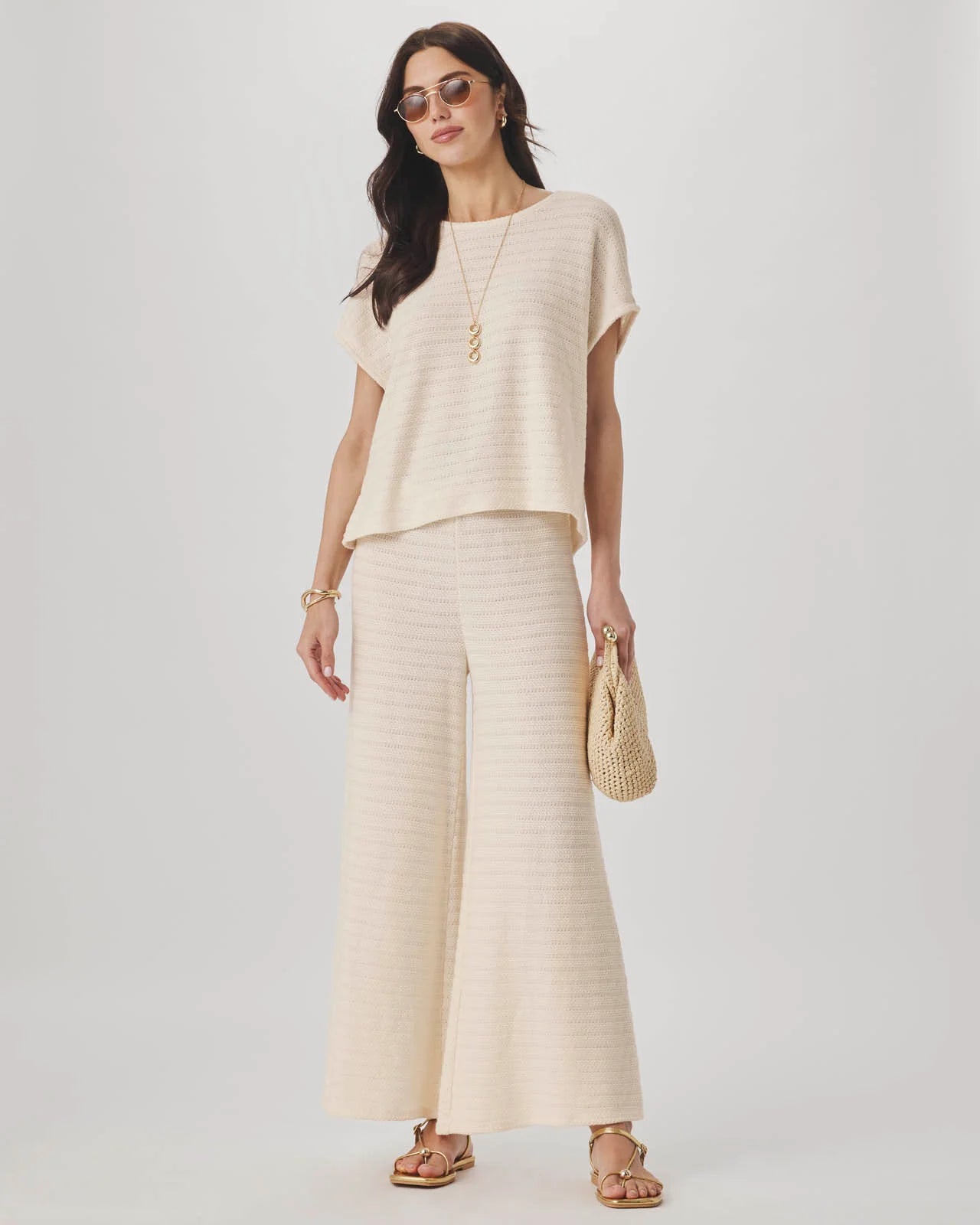 Woman wearing a beige outfit with wide-leg pants and a top, holding a straw bag on a light gray background.