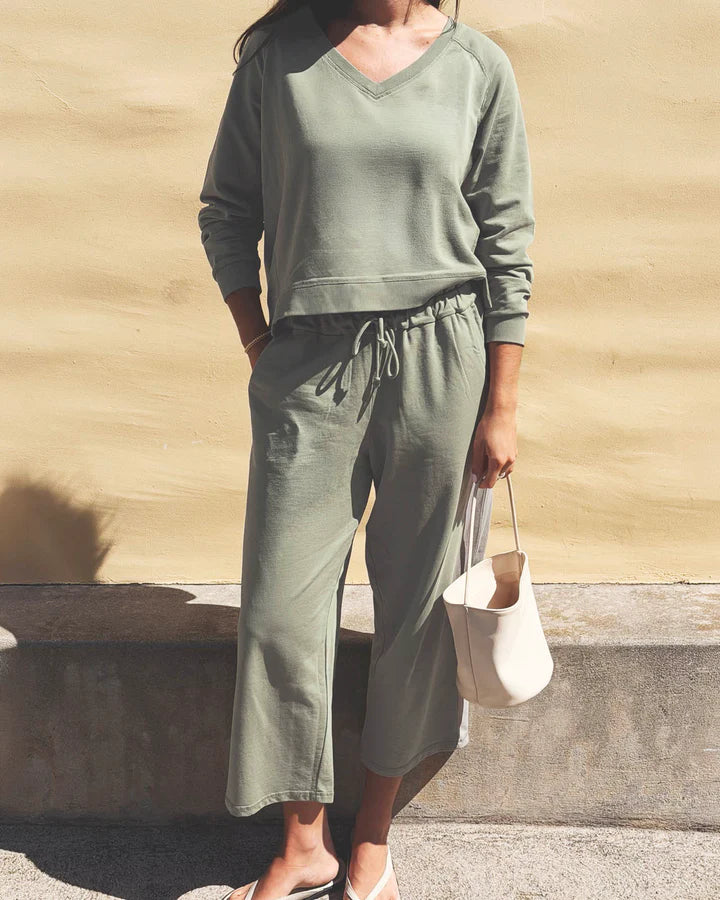 A woman wearing a matching sage green lounge set consisting of a long-sleeve V-neck top and drawstring cropped pants, holding a cream-colored tote bag against a textured beige wall.
