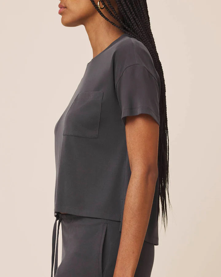 Side profile of a model wearing a dark gray short-sleeve crop top with a chest pocket, paired with matching dark gray bottoms against a plain beige background.