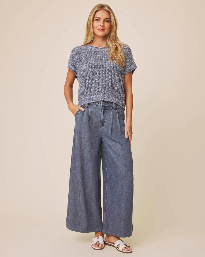 A woman modeling a short-sleeve textured knit top and wide-leg denim pants against a plain beige studio background.