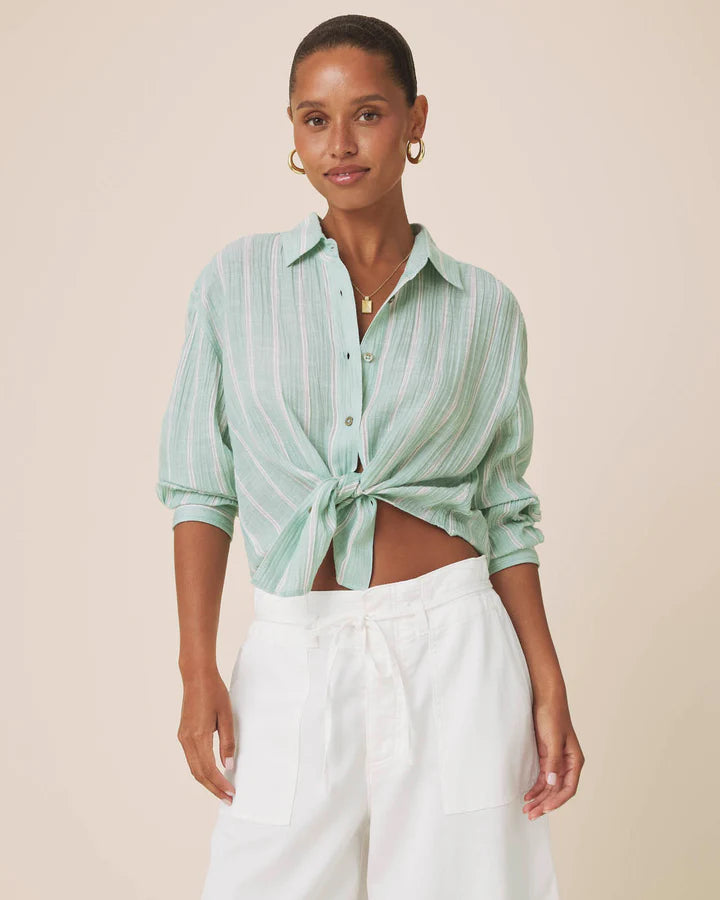 A woman modeling a mint green and white vertically striped button-down shirt tied at the waist, paired with white trousers, against a solid beige background.