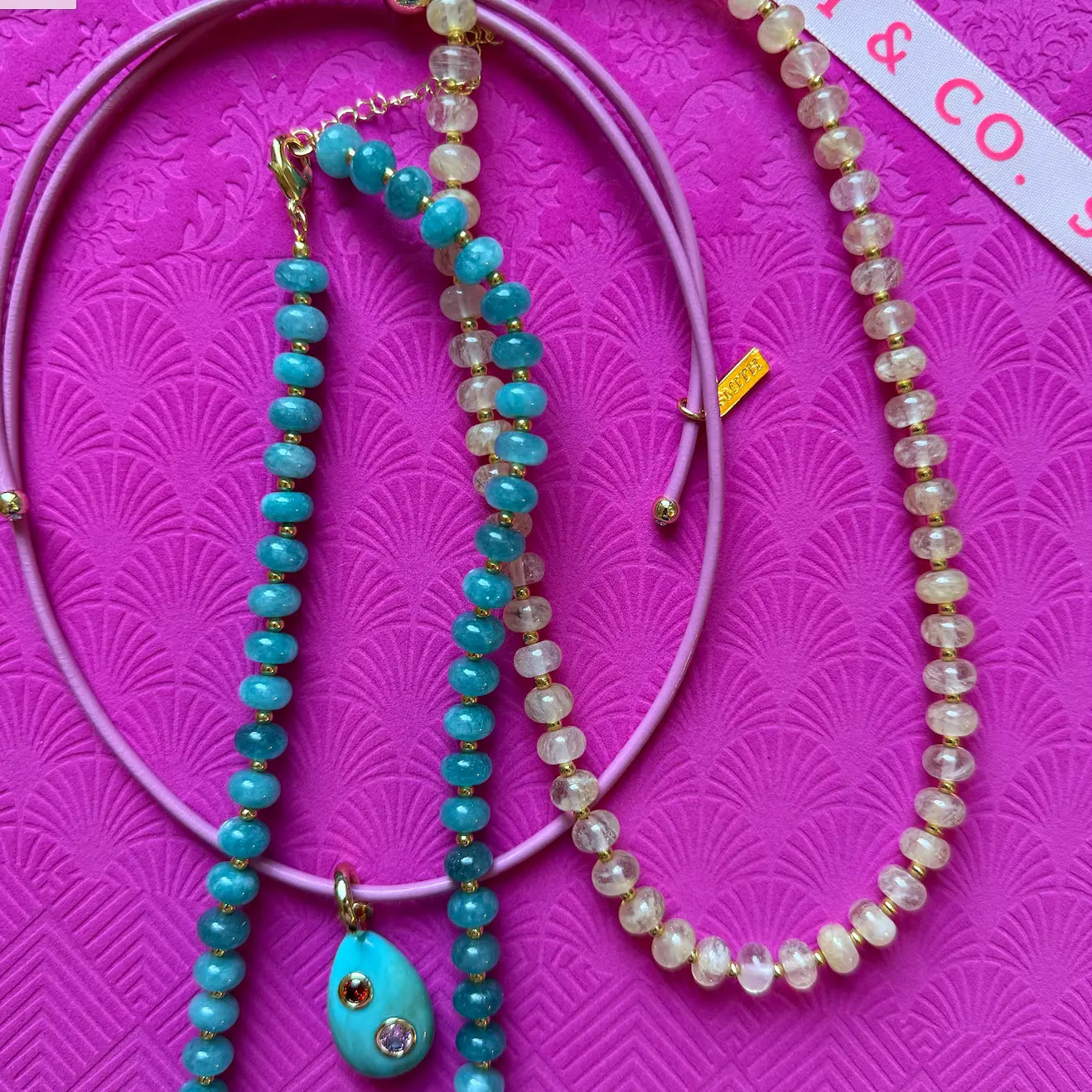 A jewelry flat-lay featuring a lavender purple beaded necklace with a turquoise teardrop pendant, a matching turquoise and clear bead strand, and a larger clear bead necklace, all arranged on a textured hot pink background.