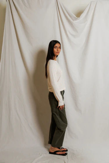Person wearing a white sweater and green pants standing against a plain white curtain.