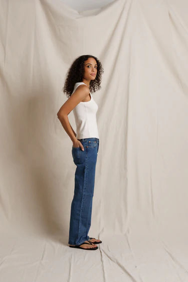 Woman wearing a white tank top and blue jeans standing against a plain background