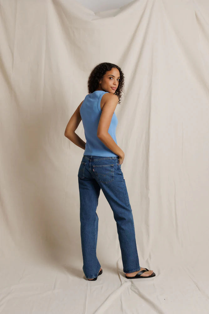 Woman wearing a blue sleeveless top and jeans standing against a plain background