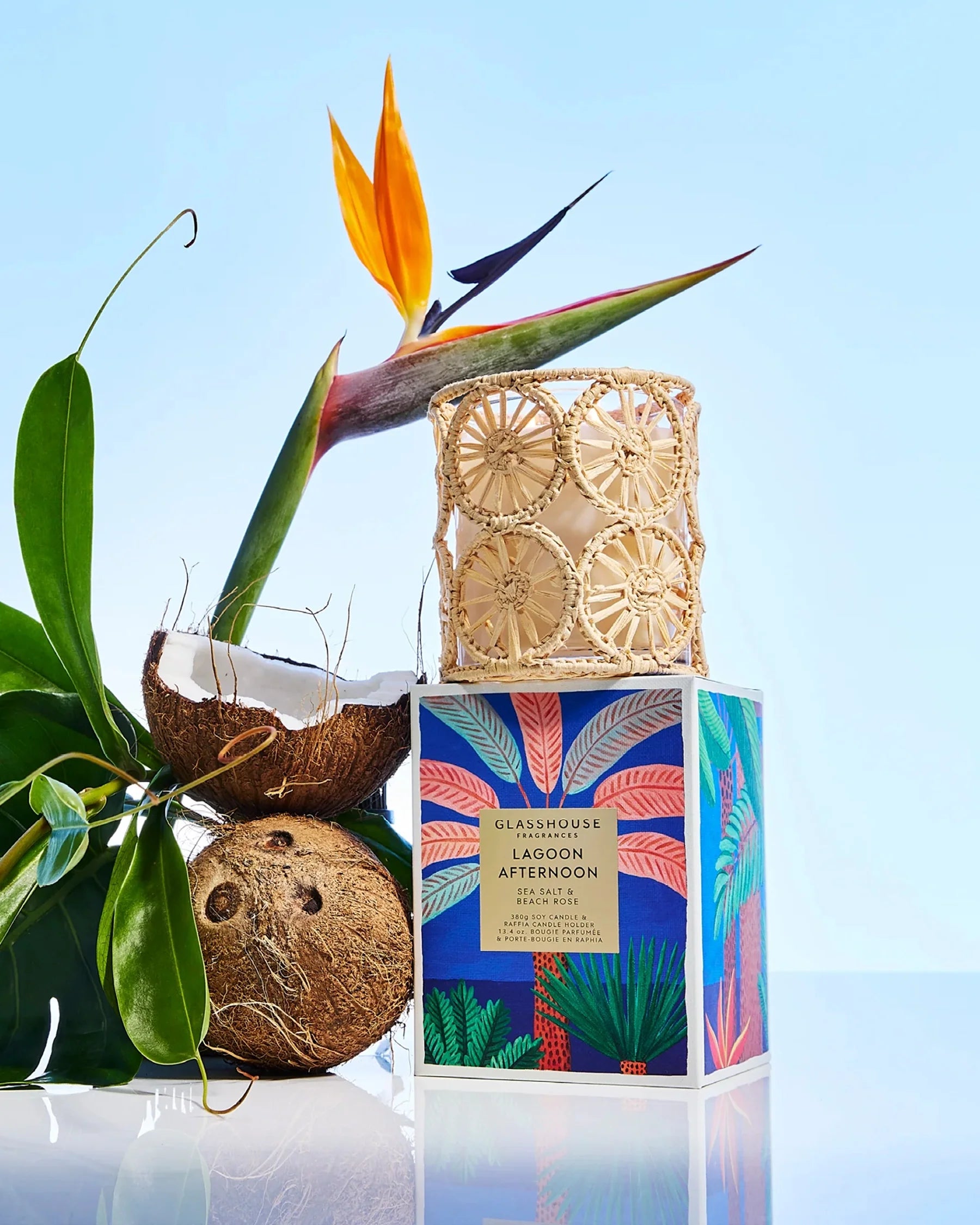 Candle with decorative packaging featuring a coconut and bird of paradise plant on a reflective surface.