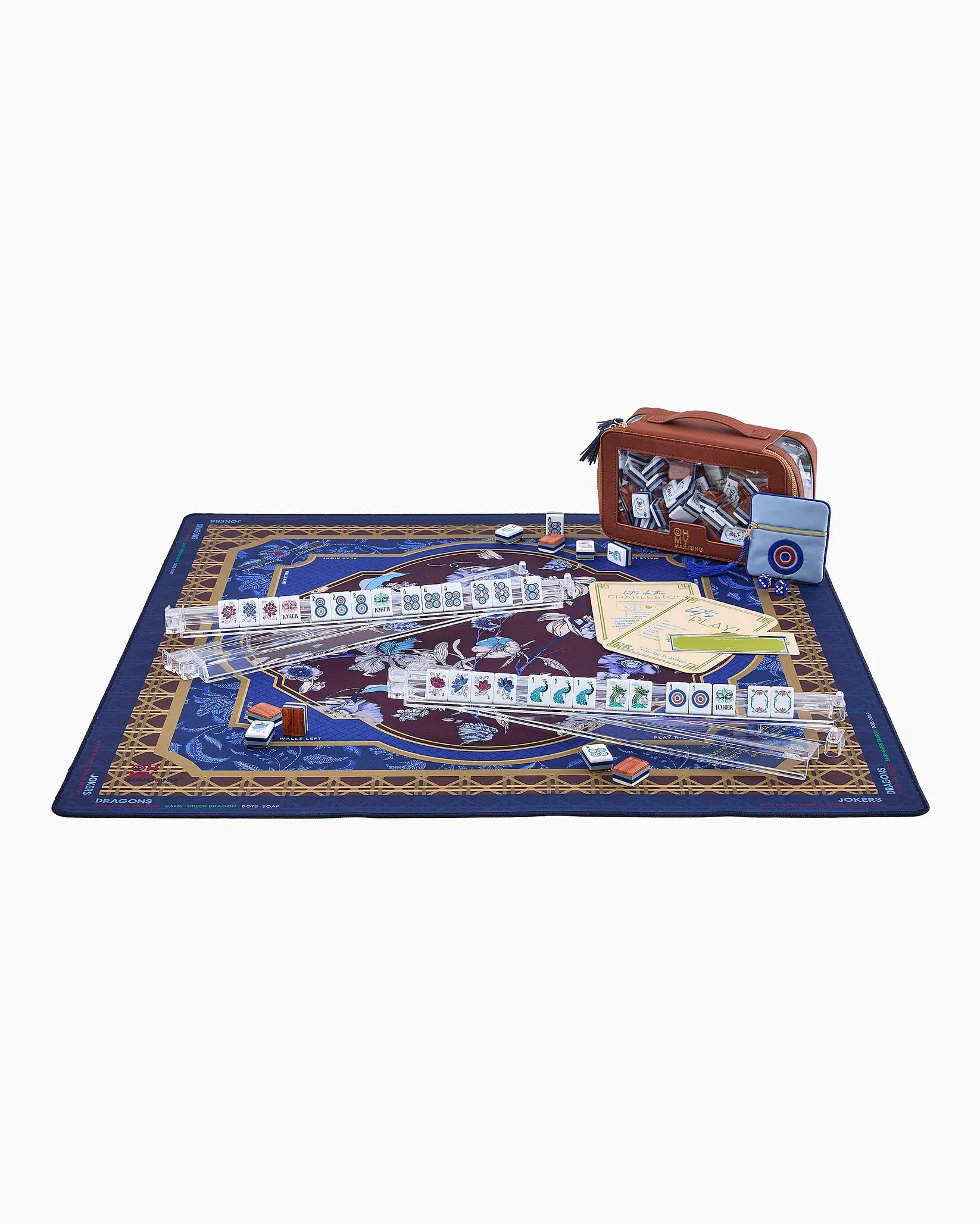 A mahjong set displayed on a decorative blue and gold mat, with several clear acrylic racks holding tiles, a brown carrying case, and game accessories arranged on a white background.