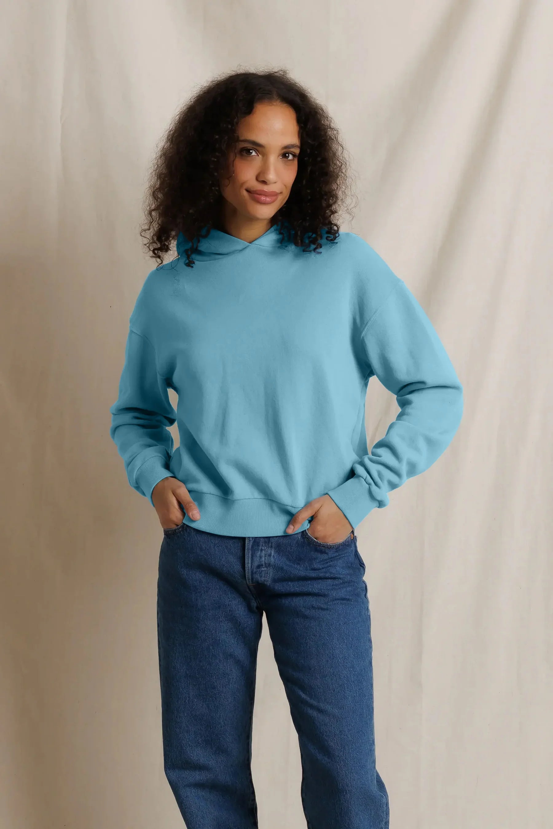 A woman with curly dark hair wearing a light blue long-sleeve mock-neck sweatshirt and blue jeans, standing against a draped beige fabric backdrop.