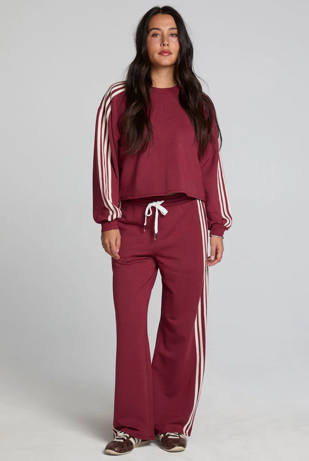 A woman modeling a matching burgundy tracksuit with white stripes on the sleeves and pants, featuring a crewneck sweatshirt and wide-leg sweatpants with a white drawstring, set against a plain light gray studio background.