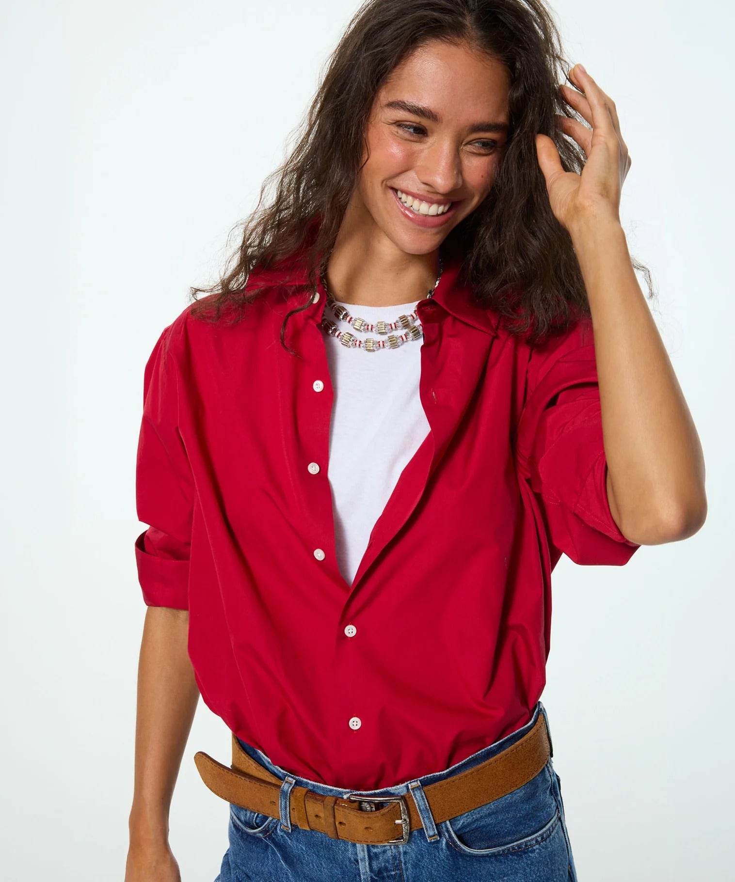 A woman wearing a bright red button-up shirt over a white t-shirt, paired with blue jeans and a brown belt, posing against a light gray studio background.