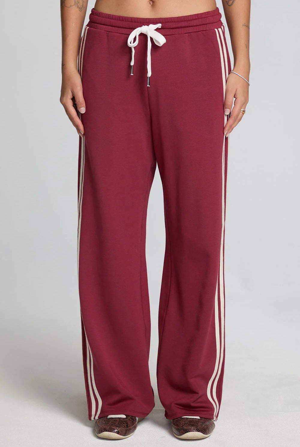 A person wearing burgundy wide-leg sweatpants with white side stripes and a white drawstring, shown from the waist down against a light gray studio background.