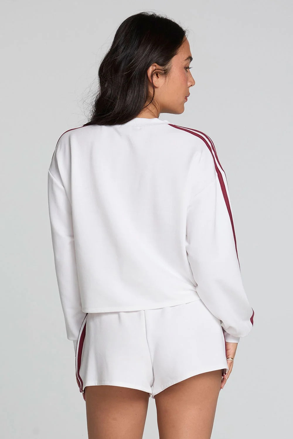 A woman shown from the back wearing a white cropped sweatshirt and matching white shorts, both with red stripes down the sleeves and sides, against a light gray studio background.